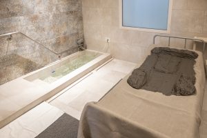 Cabina per trattamenti termali con lettino per fangoterapia, fanghi e vasca di acqua termale in primo piano.