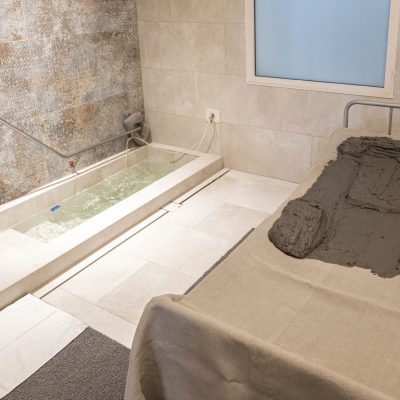 Cabina per trattamenti termali con lettino per fangoterapia, fanghi e vasca di acqua termale in primo piano.